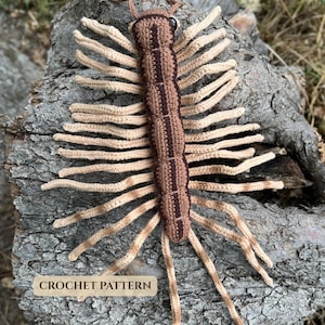 Peut inclure: Un mille-pattes en crochet avec un corps segmenté dans des tons de brun clair et foncé, doté de nombreuses longues pattes beige clair de chaque côté. Le jouet possède un petit œil noir et une étiquette en bas indiquant CROCHET PATTERN.