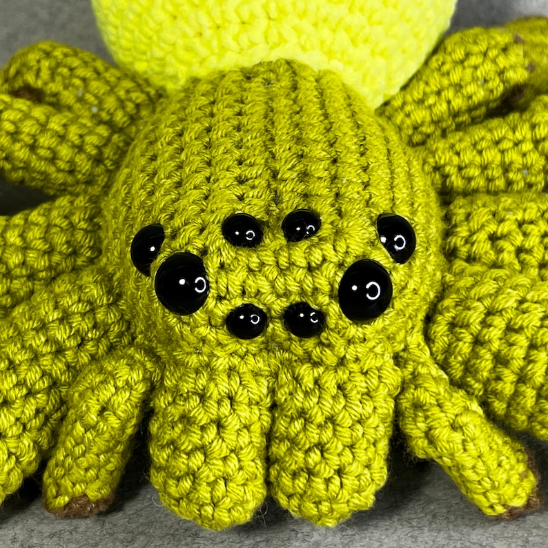 May include: A crocheted green spider with black eyes. The spider has eight legs and a round body.