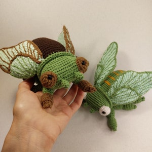 May include: Two crocheted insect toys. One is a green beetle with brown legs and wings. The other is a green cicada with brown stripes on its wings.