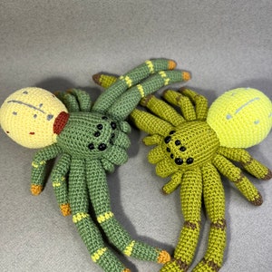 May include: Two crocheted spider plushies, one green with yellow accents and a yellow head with red markings, the other green with yellow accents and a yellow head.