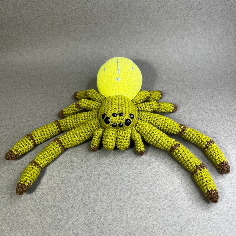 May include: A crocheted green spider with a yellow head and black eyes. The spider has eight legs and is made of yarn.