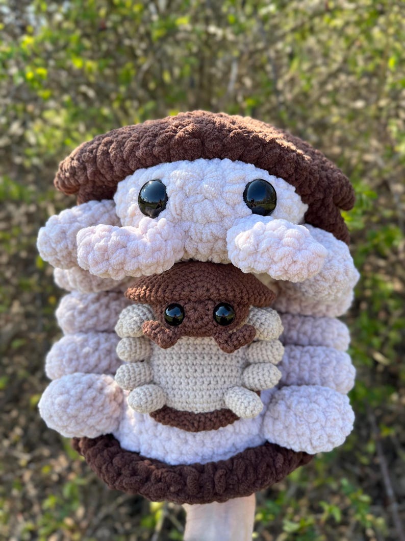 May include: A large, round, crocheted plush toy featuring a brown top and bottom with a light pink, textured, layered middle section. It has two large black plastic eyes. A smaller, brown and cream-coloured crocheted creature is attached to the front.