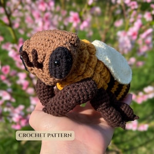May include: A crocheted bee toy featuring a brown head with black eyes, a golden yellow and brown striped body, and white wings. The toy has dark brown legs and antennae. The text CROCHET PATTERN is visible at the bottom of the image.