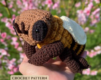 May include: A crocheted bee toy featuring a brown head with black eyes, a golden yellow and brown striped body, and white wings. The toy has dark brown legs and antennae. The text CROCHET PATTERN is visible at the bottom of the image.