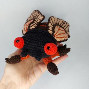 May include: A crocheted stuffed toy cicada with brown and orange legs, black body, and large red eyes with black pupils. The wings are brown and tan with a detailed crocheted pattern.