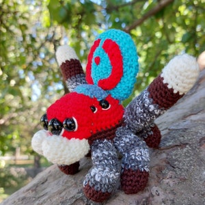 Puede incluir: Un juguete de ara&ntilde;a amigurumi tejido a crochet con un esquema de colores rojo, blanco, azul y gris. La ara&ntilde;a tiene ojos grandes y redondos y un dise&ntilde;o &uacute;nico y colorido.