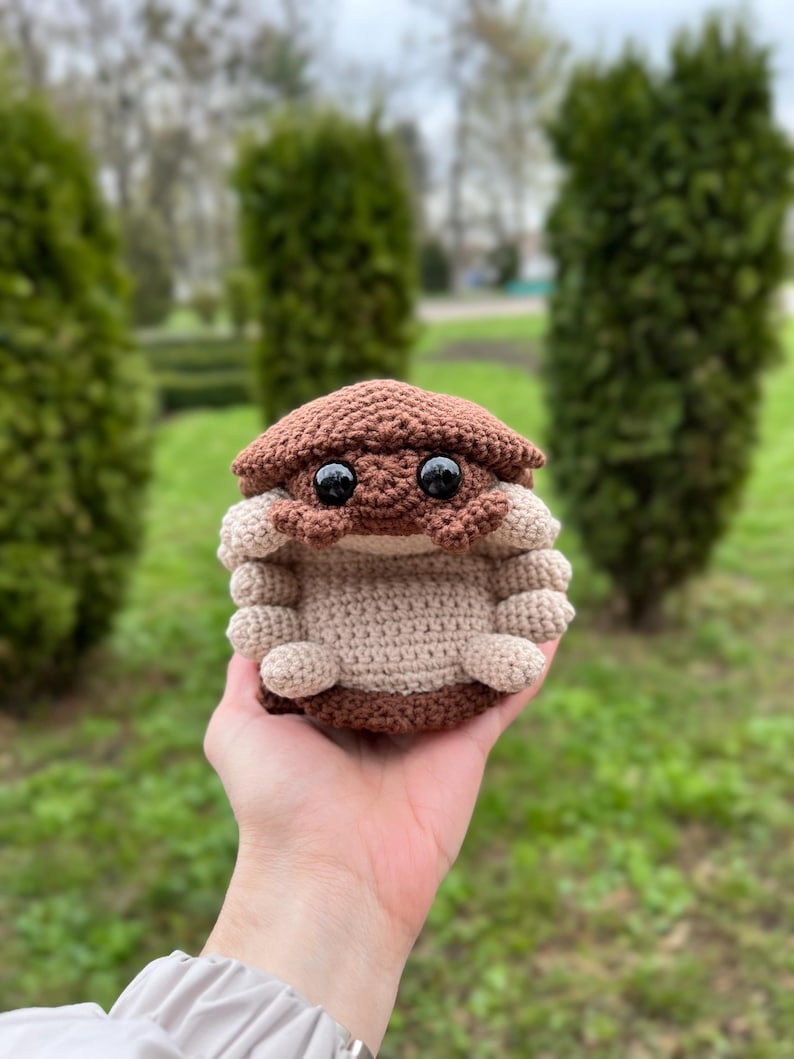 May include: A small, round, crocheted plush toy shaped like an isopod. The creature features a dark brown segmented shell on top, a light beige underside, and multiple small, rounded beige legs along the sides. It has two large, glossy black eyes.