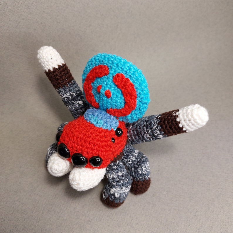 Puede incluir: Un juguete de ara&ntilde;a de crochet con un cuerpo rojo, patas blancas y una espalda con un patr&oacute;n azul y rojo. La ara&ntilde;a tiene ojos negros y una cara blanca.
