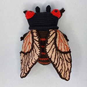 May include: A crocheted cicada toy with brown and beige wings, red eyes, and a black head.