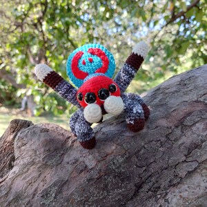 Puede incluir: Un juguete de ara&ntilde;a de crochet con un esquema de colores azul, rojo y blanco. La ara&ntilde;a tiene ojos grandes y negros y un cuerpo rojo.