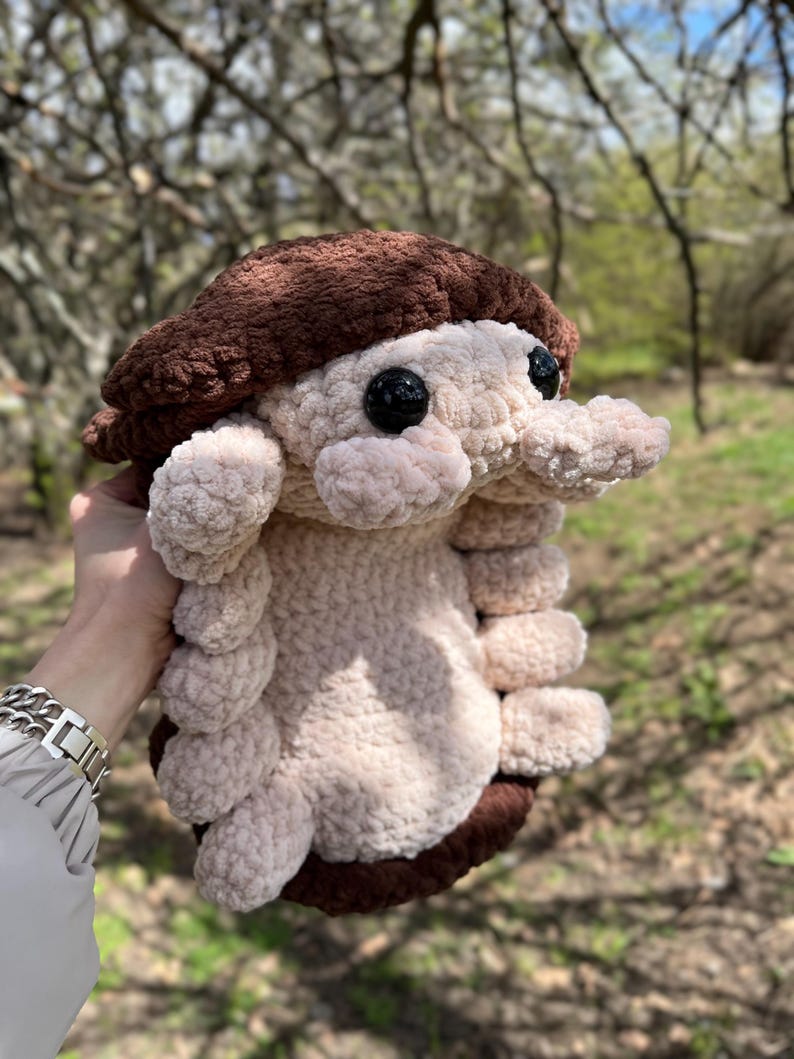 May include: A plush toy shaped like an isopod, featuring a dark brown textured cap and a light beige segmented body. The creature has two large, round, black plastic eyes and multiple small, soft, rounded legs extending from its sides.