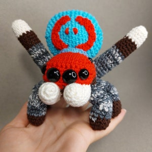 Puede incluir: Una ara&ntilde;a de juguete de crochet con una cabeza con estampado rojo y azul, ojos negros y patas grises y marrones.