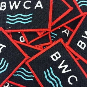 May include: A close-up of several black fabric patches with red stitching. Each patch features the text "BWCA" in white with a blue wave design.