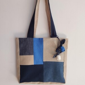 Könnte beinhalten: Eine rechteckige Tragetasche mit einem Patchwork-Design aus Jeans- und Canvas-Stoff in Marineblau, Hellblau und Beige. Die Tasche verfügt über zwei Schulterriemen und einen dekorativen Anhänger an der Seite, der aus kleinen Stoffknoten besteht.