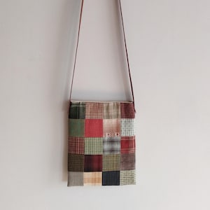Może przedstawiać: Torba na ramię z patchworku z brązowym skórzanym paskiem. Korpus torby składa się z różnych kwadratów materiału w odcieniach czerwieni, zieleni, brązu i beżu. Torba wisi na gładkiej białej ścianie.