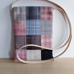 Könnte beinhalten: Eine Patchwork-Umhängetasche mit einem hellbraunen Lederriemen. Die Tasche besteht aus verschiedenen Stoffquadraten in Rosa-, Blau-, Grau- und Schwarztönen. Die Tasche liegt auf einer Holzoberfläche.