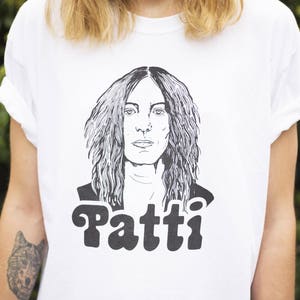Pu&ograve; includere: Maglietta bianca con un'illustrazione in bianco e nero di Patti Smith, cantante e cantautrice, con il testo "Patti" stampato sotto l'illustrazione.