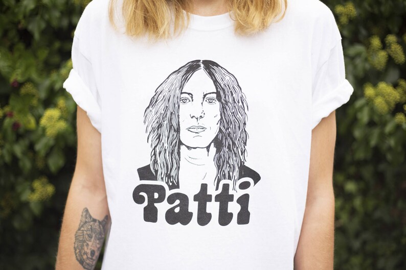 Pu&ograve; includere: Maglietta bianca con un'illustrazione in bianco e nero di Patti Smith, cantante e cantautrice, con il testo "Patti" stampato sotto l'illustrazione.