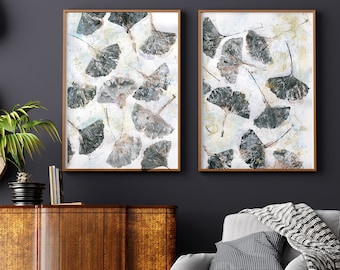 May include: Two framed art prints featuring ginkgo leaf designs. The leaves are in shades of gray and green, set against a white and cream-colored background. The frames are a warm brown color, adding a natural touch to the artwork.