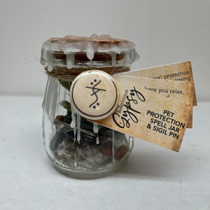 May include: A clear glass jar with a cork lid sealed with white wax. The jar contains small stones, herbs, and salt. A small tag reads "PET PROTECTION SPELL JAR & SIGIL PIN". A round pin with a symbol is attached to the jar.