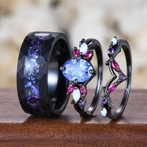 May include: A three-piece ring set featuring a wide black band with a faceted surface and a purple and blue iridescent inlay, alongside two black bands adorned with oval blue stones, small white opals, and marquise-cut pink and purple gems.