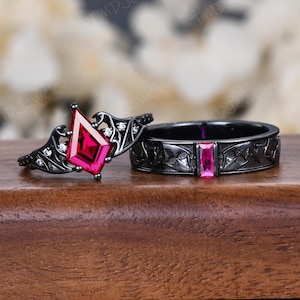 Può includere: Un set di anelli in metallo nero con una pietra rossa a forma di aquilone su un anello e una pietra rossa taglio baguette sull'altro. Entrambi gli anelli sono decorati con intricati motivi ad ala e piccole pietre trasparenti.