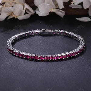 May include: A silver tennis bracelet with a row of vibrant, round, ruby-colored gemstones. The bracelet is set against a dark background, with white flowers in the background. The clasp is marked with "S925".
