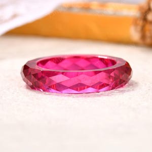 May include: A vibrant fuchsia pink ring featuring a faceted, geometric surface with multiple triangular cuts that create a sparkling, diamond-like texture. The band is uniform in width and has a polished, translucent appearance throughout the entire piece.