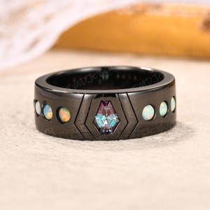 May include: A black metal ring featuring a central coffin-shaped alexandrite stone set within a geometric frame, flanked by a series of small, round, iridescent opals embedded along the band, creating a distinct contrast between the dark metal and stones.
