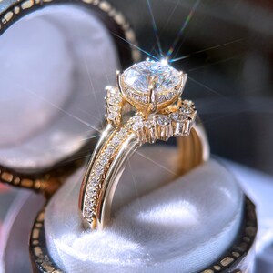 May include: A gold engagement ring with a large, clear, round-cut diamond as the centerpiece. The ring features a halo of smaller diamonds around the main stone and along the band, set in a gold band. The ring is displayed in a velvet-lined ring box.