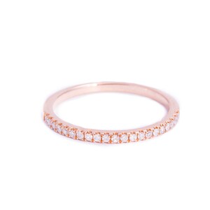 May include: A rose gold band ring featuring a continuous row of small, clear, round-cut stones set along the front half of the band. The metal has a smooth, polished finish, and the stones are arranged in a single, uniform line across the surface.