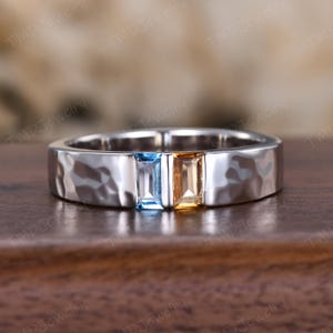 May include: A silver ring with a hammered texture features two rectangular gemstones: a light blue and a golden-yellow. The ring is set on a wooden surface, with the TMD Jewelry logo visible in the background.