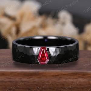 Puede incluir: Un anillo negro con textura martillada y una vibrante gema roja. La gema tiene una forma geométrica única, engastada en un metal plateado o blanco. El anillo se exhibe sobre una superficie de madera.