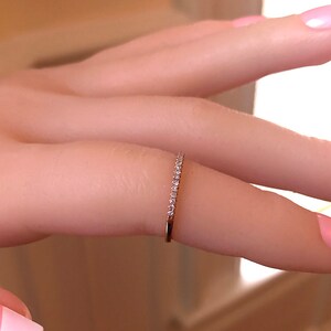 May include: A thin gold band ring featuring a row of small, clear, round-cut stones set along the top half of the band. The metal has a warm, metallic yellow tone, and the stones are arranged in a continuous line, creating a delicate, sparkling appearance.