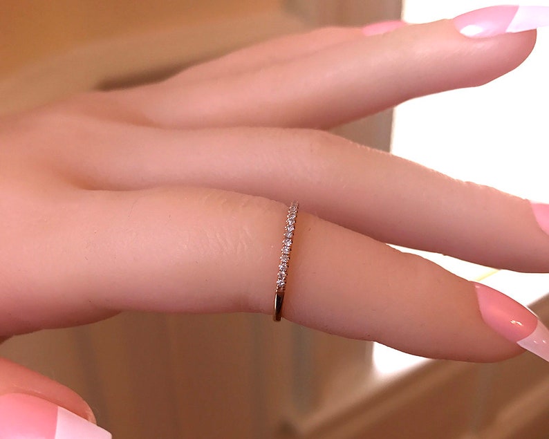 May include: A thin gold band ring featuring a row of small, clear, round-cut stones set along the top half of the band. The metal has a warm, metallic yellow tone, and the stones are arranged in a continuous line, creating a delicate, sparkling appearance.