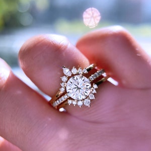 May include: A gold engagement ring set featuring a large, round, clear gemstone surrounded by smaller stones in a sunburst design. The band is also adorned with smaller gemstones. The ring is held up by a person's fingers.