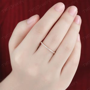 May include: A thin, rose gold band ring featuring a continuous row of small, clear, round-cut pav&eacute;-set stones along the top half of the band. The ring is worn on the ring finger, showcasing a delicate and slender design with a polished metal finish.