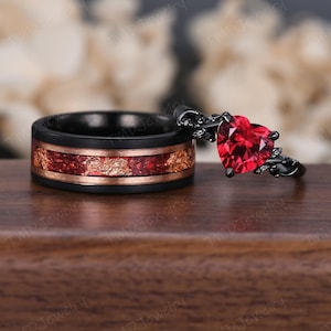 May include: A black and red wedding ring set. The man's ring is a black tungsten band with a red and copper inlay. The woman's ring is a black band with a large red heart-shaped gemstone and black floral accents.