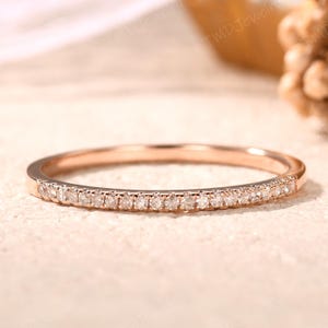 May include: A thin rose gold band ring featuring a row of small, clear, round-cut stones set along the front half of the band. The metal has a smooth, polished finish, and the stones are arranged in a continuous, uniform line across the surface.