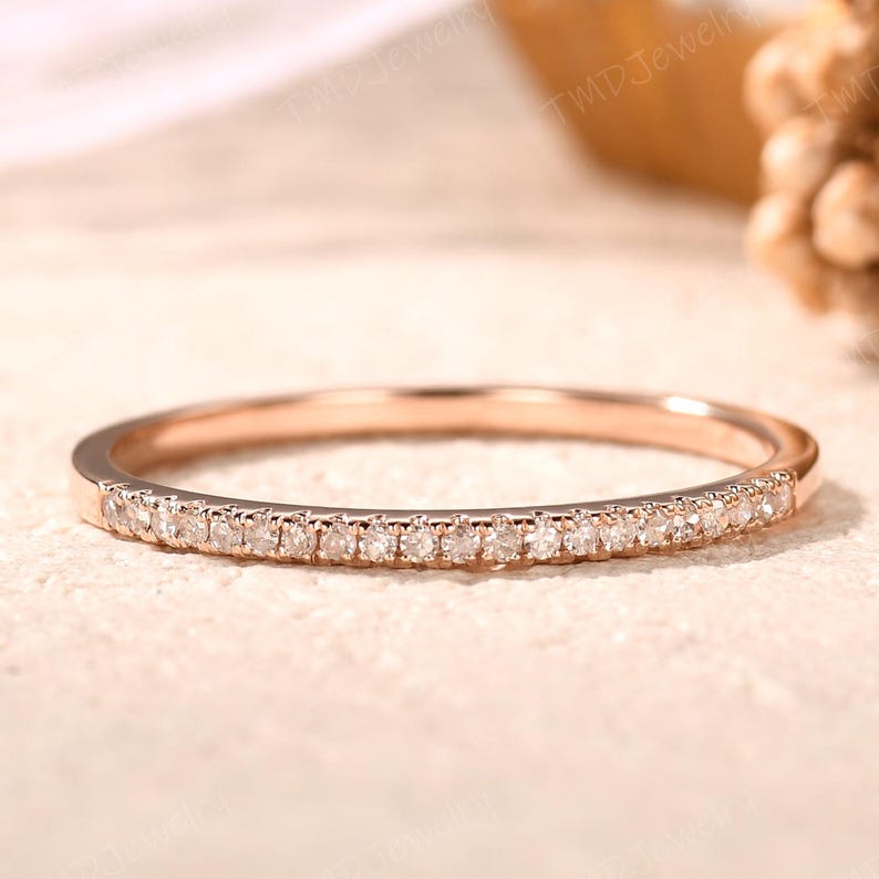 May include: A thin rose gold band ring featuring a row of small, clear, round-cut stones set along the front half of the band. The metal has a smooth, polished finish, and the stones are arranged in a continuous, uniform line across the surface.