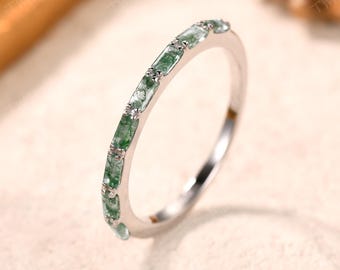 May include: A silver ring with a band of rectangular green gemstones. The gemstones are set in a row around the ring. The ring is on a light-colored surface.