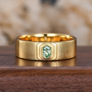May include: A gold-colored ring with a textured band and a hexagonal green stone. The ring has a brushed finish and a geometric design around the central stone. The ring is displayed on a wooden surface.