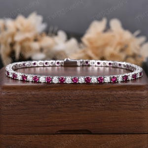 May include: A silver bracelet featuring alternating round-cut, vibrant red gemstones and clear, sparkling stones. The bracelet is displayed on a wooden surface, showcasing its elegant design and craftsmanship. The clasp is visible, highlighting the secure closure.