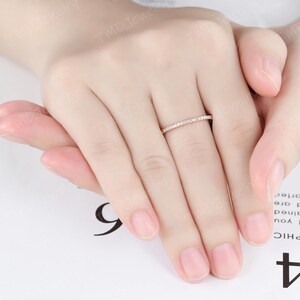 May include: A thin rose gold band ring featuring a continuous row of small, clear, round-cut pav&eacute;-set stones along the top half of the band. The ring is worn on the middle finger, showcasing a delicate and slender design with a polished metal finish.