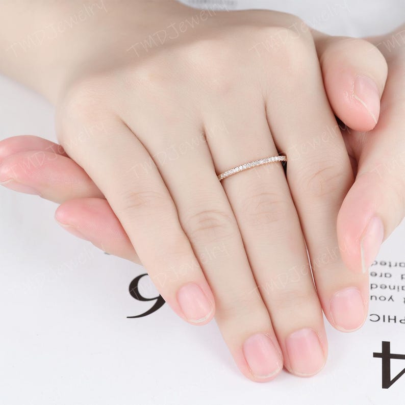 May include: A thin rose gold band ring featuring a continuous row of small, clear, round-cut pav&eacute;-set stones along the top half of the band. The ring is worn on the middle finger, showcasing a delicate and slender design with a polished metal finish.