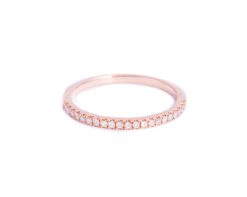 May include: A rose gold band ring featuring a continuous row of small, clear, round-cut stones set along the front half of the band. The metal has a smooth, polished finish, and the stones are arranged in a single, uniform line across the surface.