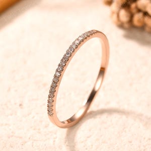 May include: A thin rose gold band ring featuring a row of small, clear, round-cut stones set along the top half of the circumference. The metal has a polished, reflective finish, and the stones are arranged in a continuous, uniform line.