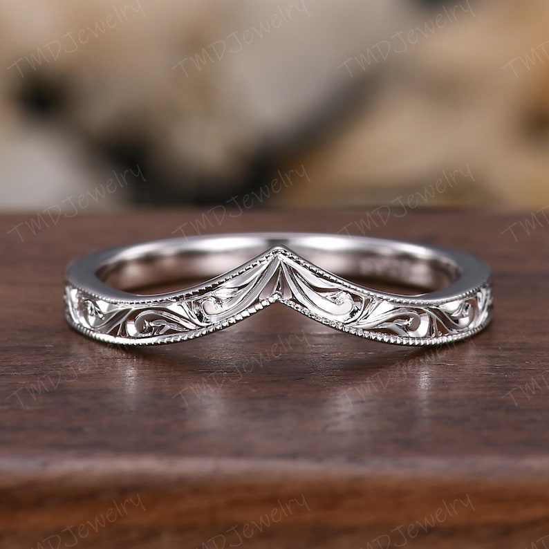 May include: A silver-toned V-shaped chevron ring featuring intricate engraved scrollwork patterns along the band. The edges of the ring are detailed with a fine milgrain texture, creating a delicate and structured appearance throughout the metal surface.