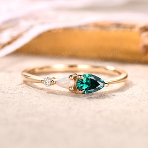 May include: A gold ring featuring three gemstones: a small diamond, an oval opal, and a pear-shaped emerald. The ring's band is thin and polished, with the gemstones set in a row.