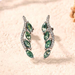 May include: A pair of silver earrings featuring a leaf design. Each earring has a curved stem adorned with small, clear stones and a row of green, oval-shaped gemstones. The earrings are set against a neutral background.